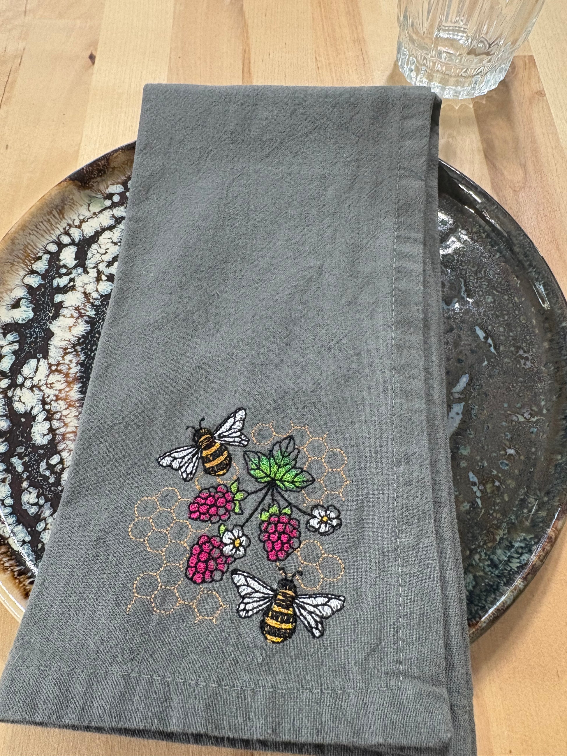 Gray embroidered napkin with bee and raspberry design on a wooden surface