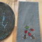 Gray towel with embroidered bees and strawberries on a wooden surface next to a glass and plate.