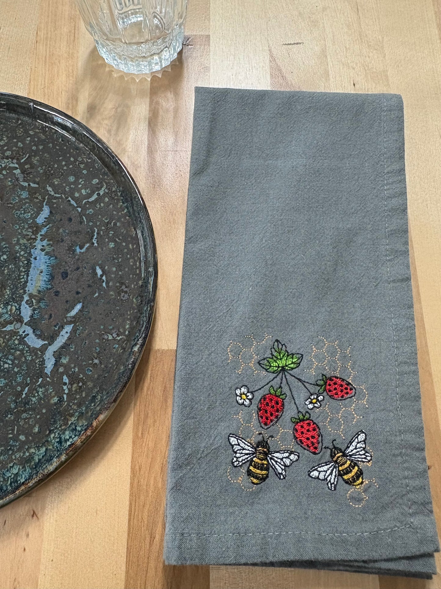 Gray towel with embroidered bees and strawberries on a wooden surface next to a glass and plate.