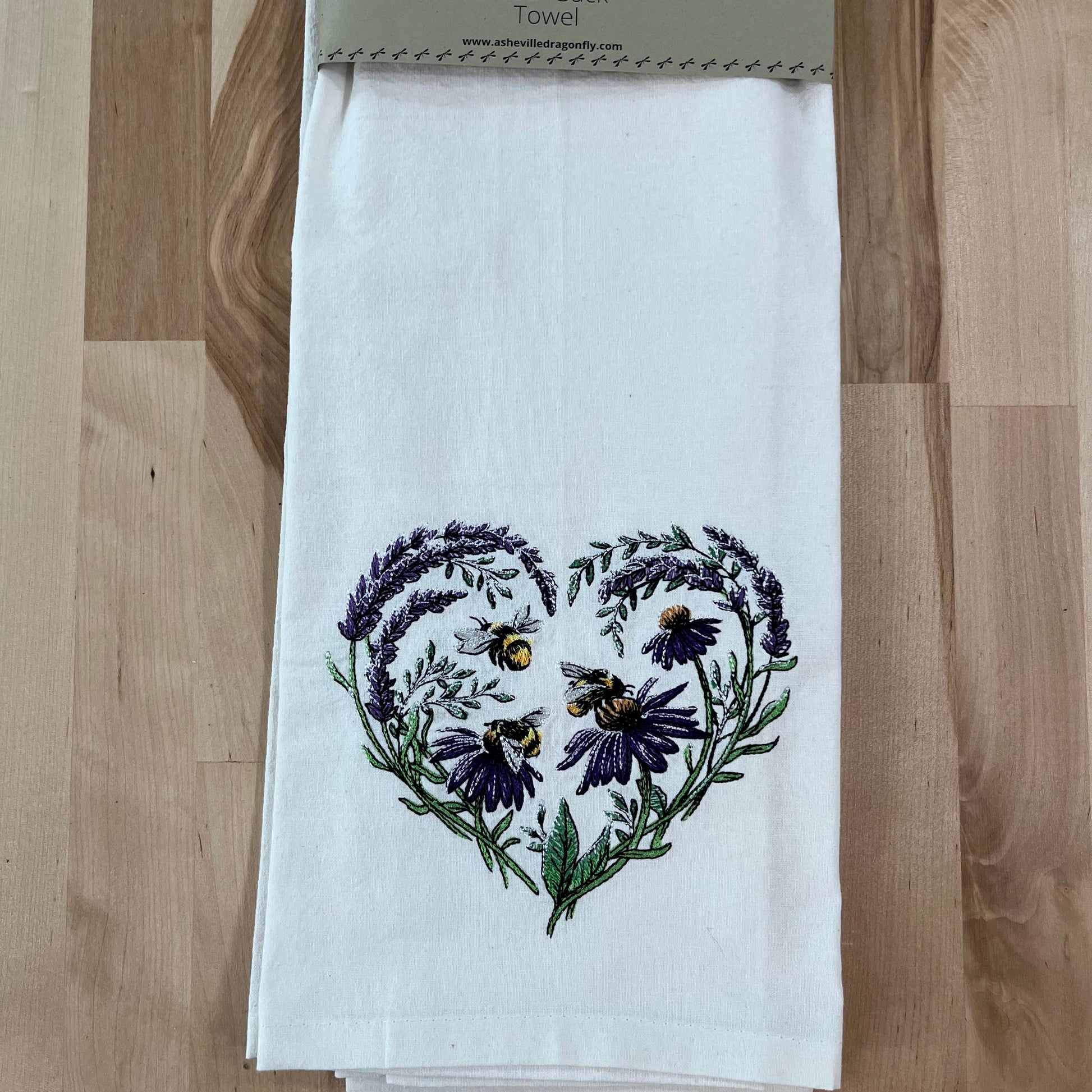 A white kitchen towel with an embroidered heart shape featuring buzzing blossoms and bees, placed on a wooden surface.