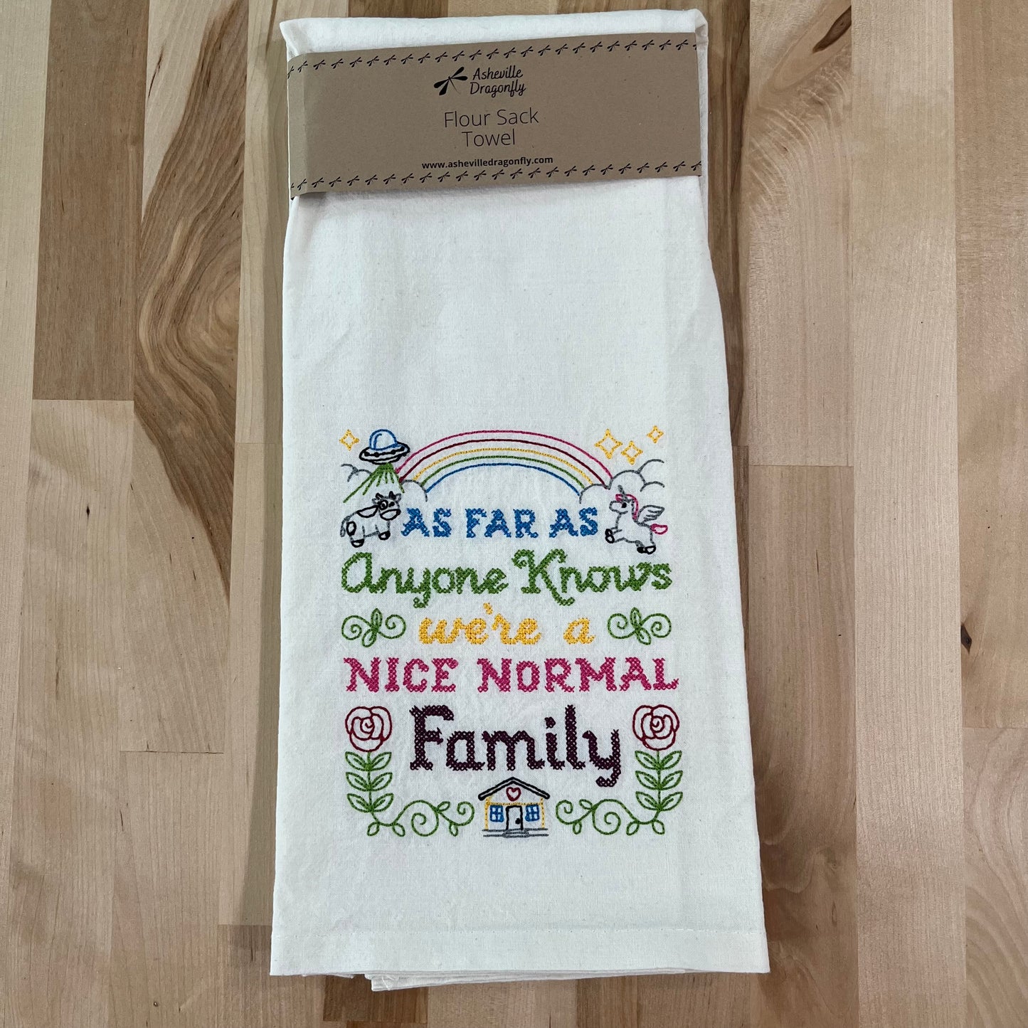 A white kitchen towel with colorful embroidered text and graphics, including a house and a rainbow, with a humorous phrase about a nice normal family.