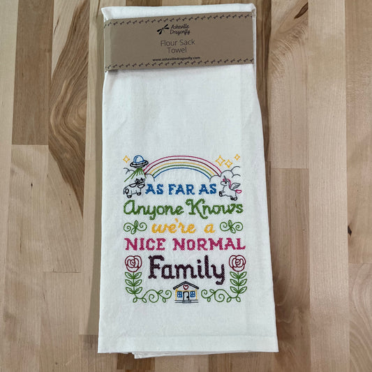 A white kitchen towel with colorful embroidered text and graphics, including a house and a rainbow, with a humorous phrase about a nice normal family.