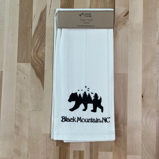 A white kitchen towel with black embroidered bear and forest design, displayed on a wooden surface with a cardboard hanger.