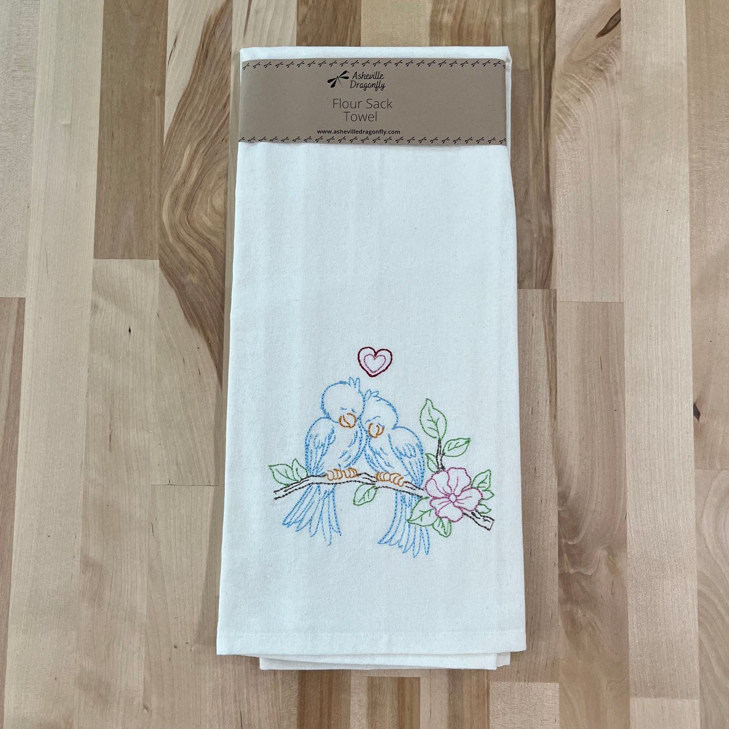 A white kitchen towel with an embroidered design of love birds perched on a branch, surrounded by a floral pattern, on a wooden surface.